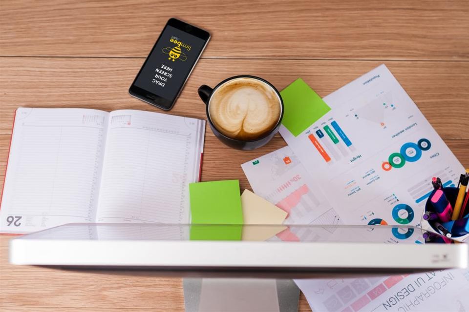 Holztisch von oben mit Notizbuch, Kaffeetasse, Smartphone, Haftzetteln, Diagrammen und Monitorrand, Symbolbild