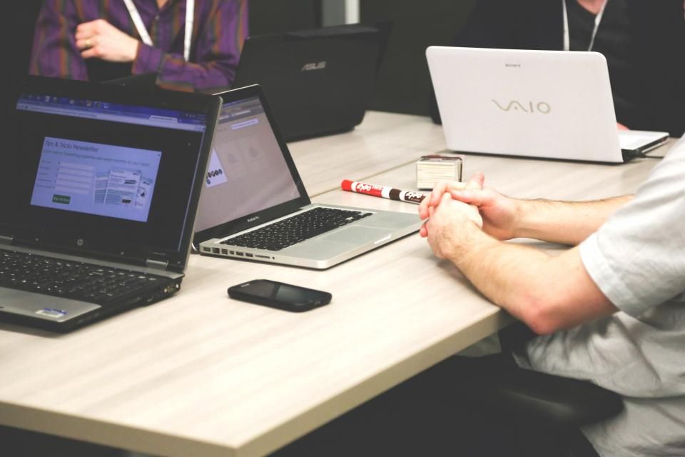 Auf einem langen Tisch stehen mehrere Laptops, eine Person sitzt mit gefalteten Händen davor, Symbolbild