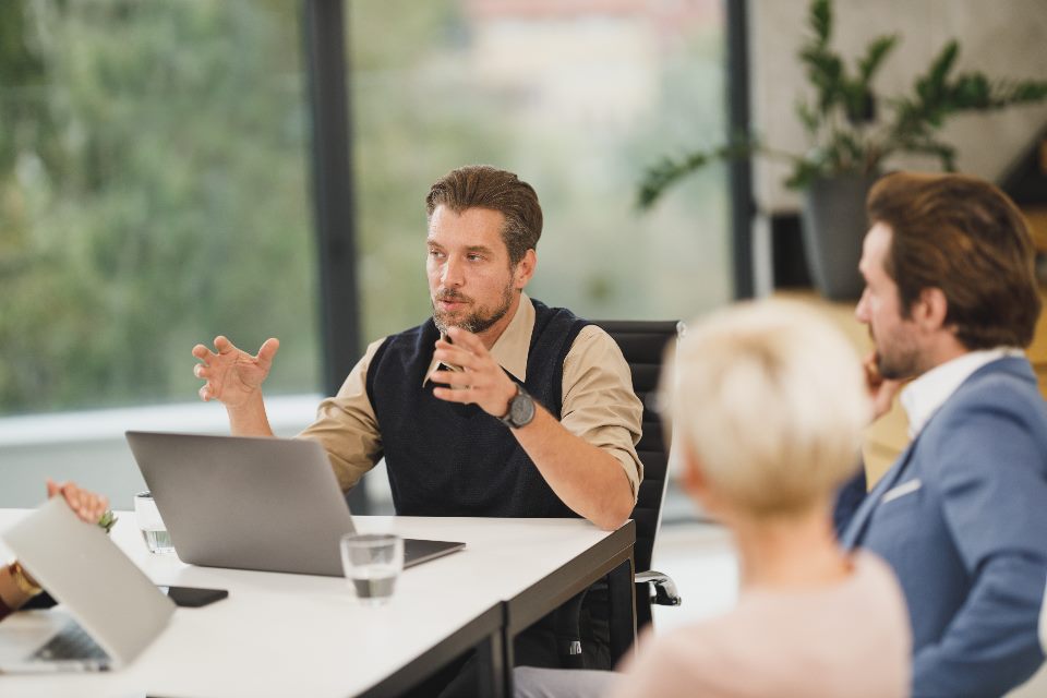 Mann im Strickpullunder gestikuliert am Besprechungstisch mit Laptops und Wassergläsern vor weiteren Personen, Symbolbild