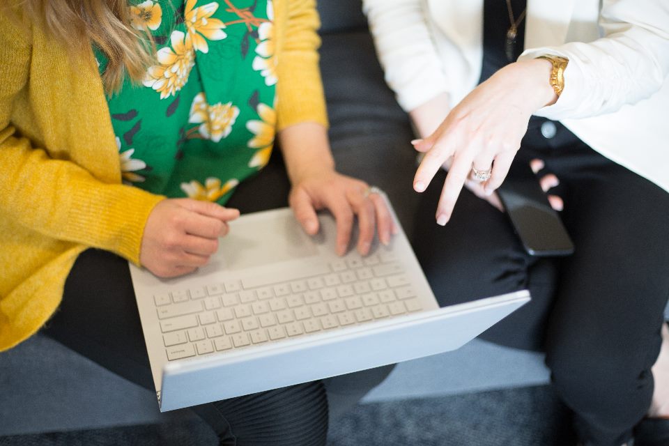 Zwei Personen sitzen mit einem Laptop auf dem Schoß, eine zeigt darauf, ein Smartphone liegt daneben, Symbolbild
