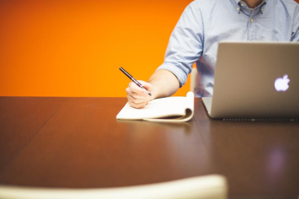 Eine Person im hellblauen Hemd schreibt mit Stift in ein Notizbuch neben einem geschlossenen Laptop vor oranger Wand, Symbolbild