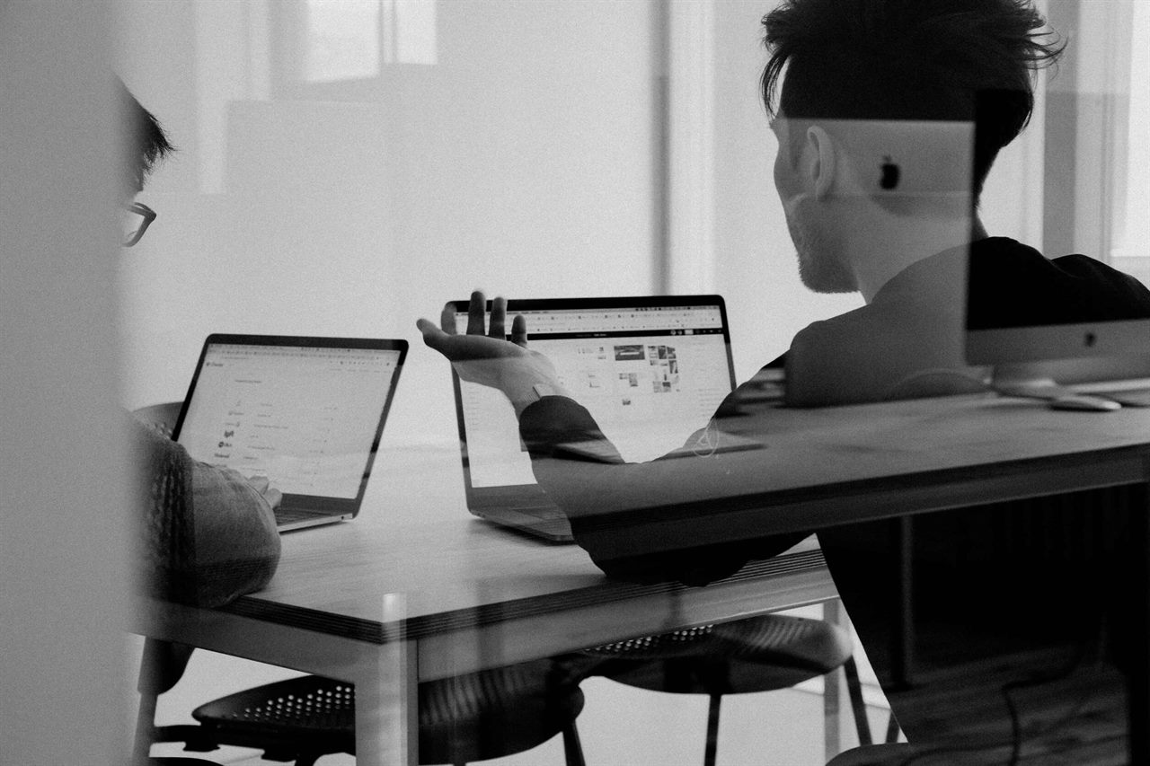 Zwei Personen an Laptops in einem schwarz-weißen Büro, durch eine Glasscheibe fotografiert